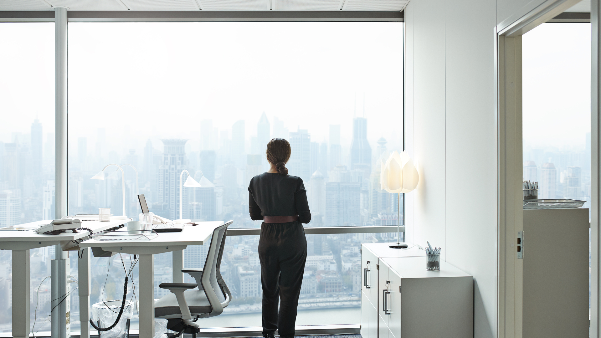 Businesswoman standing inside office and looking out of big window with beautiful view of skyline