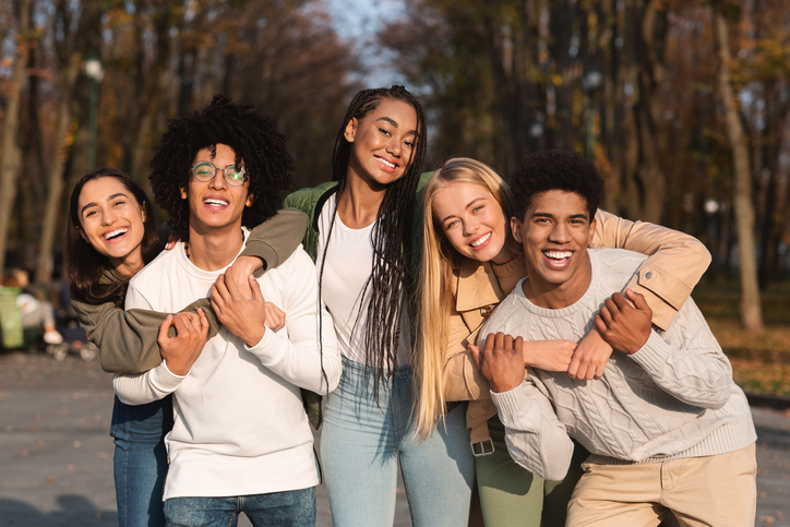 a group of mixed race teenagers smiling and laughing as they hug in front of a forest backrop