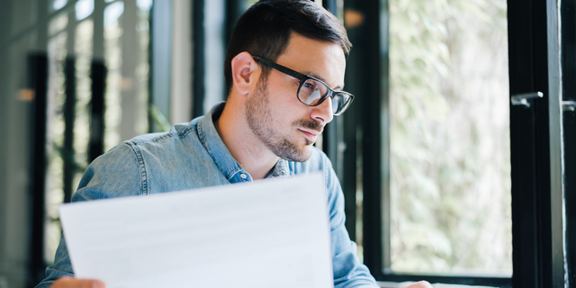 Serious pensive thoughtful focused young casual business accountant bookkeeper in office looking at and working with laptop and income tax return papers and documents