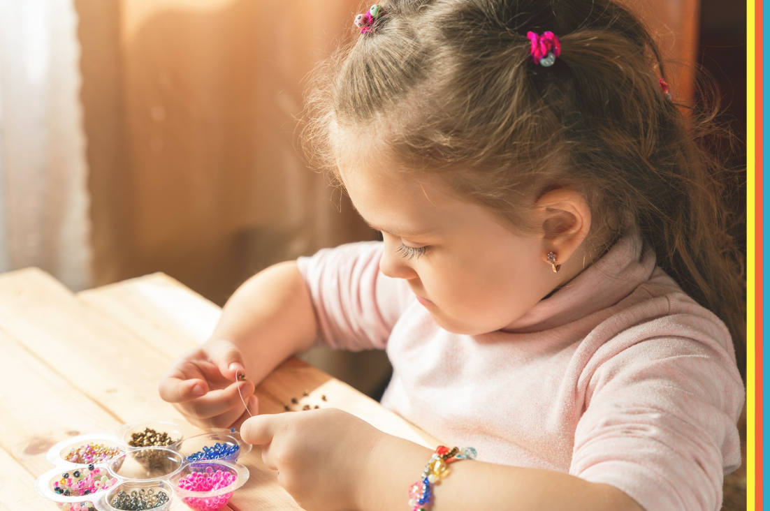 Smiling young child drawing with coloured pencils at a table in a shopping centre play area, with a blurred family-friendly background and colourful pencils in the foreground