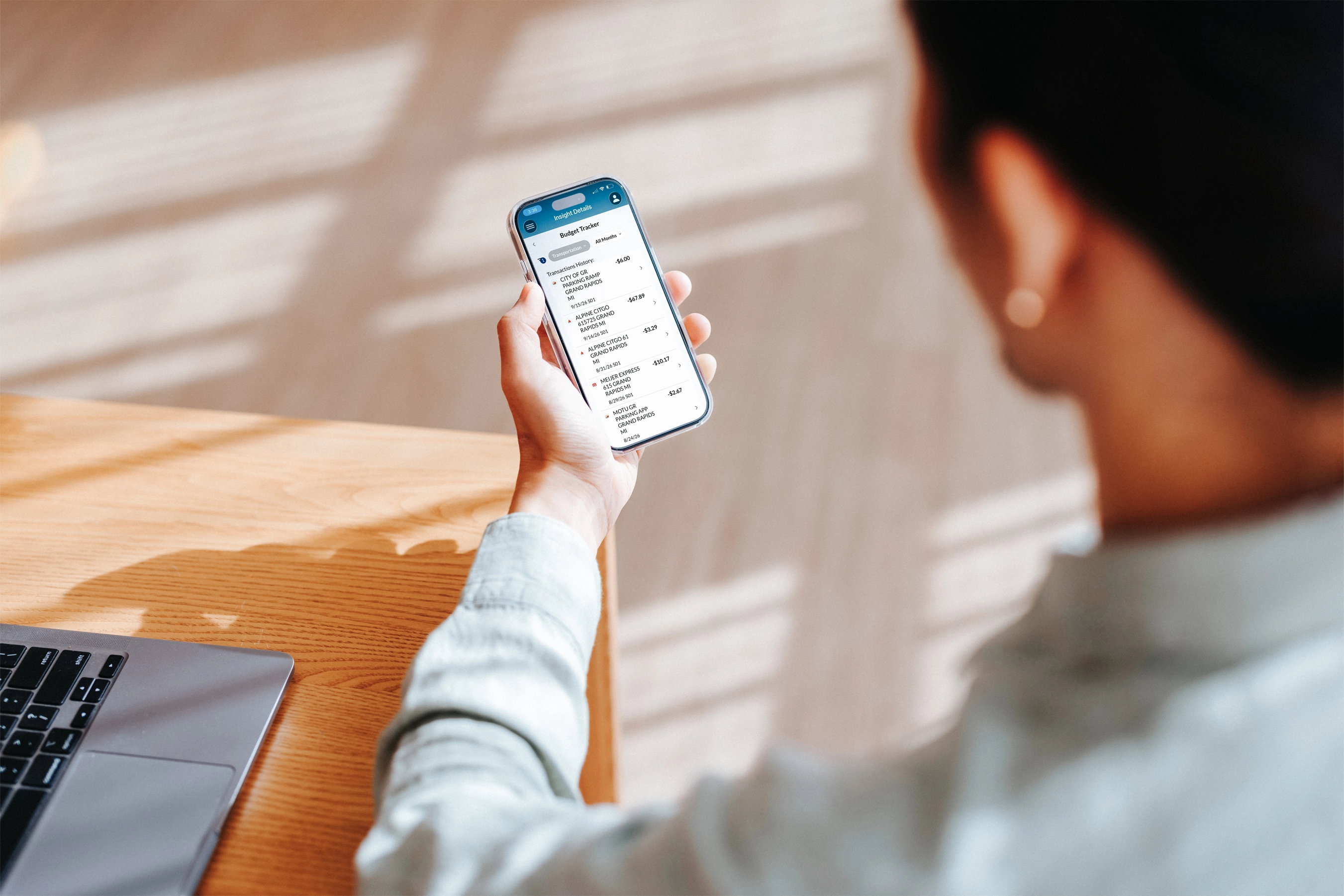 Person viewing recent transactions and budget details on a smartphone while seated at a desk with a laptop.