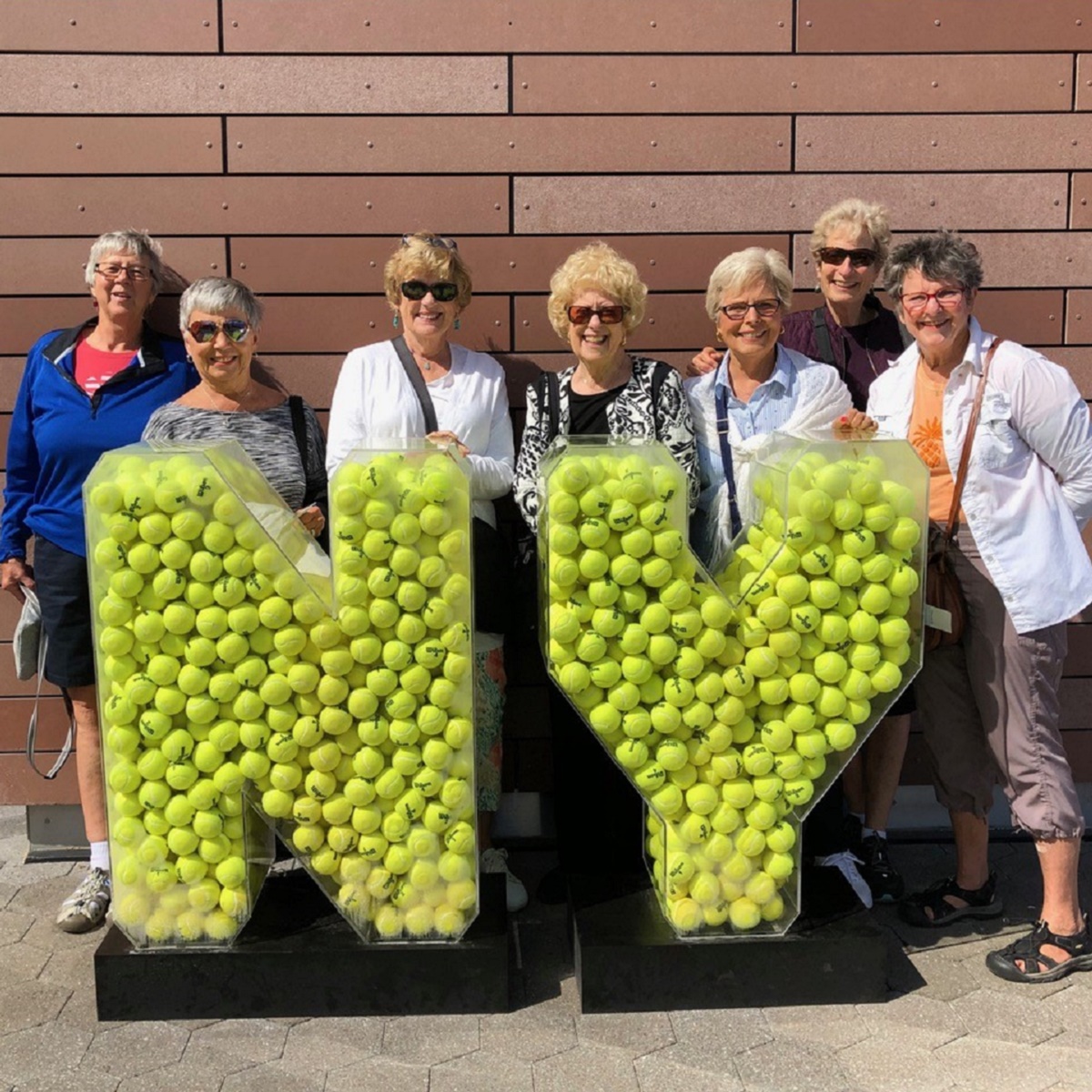 The women stand behind a statue made of tennis balls that forms the shapes of the letters "NY"