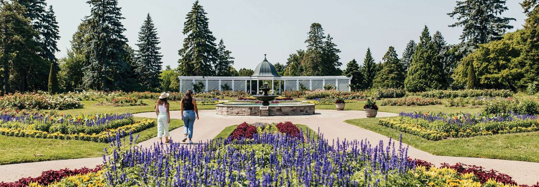 Two people walking through a lush flower garden with a stone fountain and white pavilion at the Shaw Festival in Niagara, Ontario.