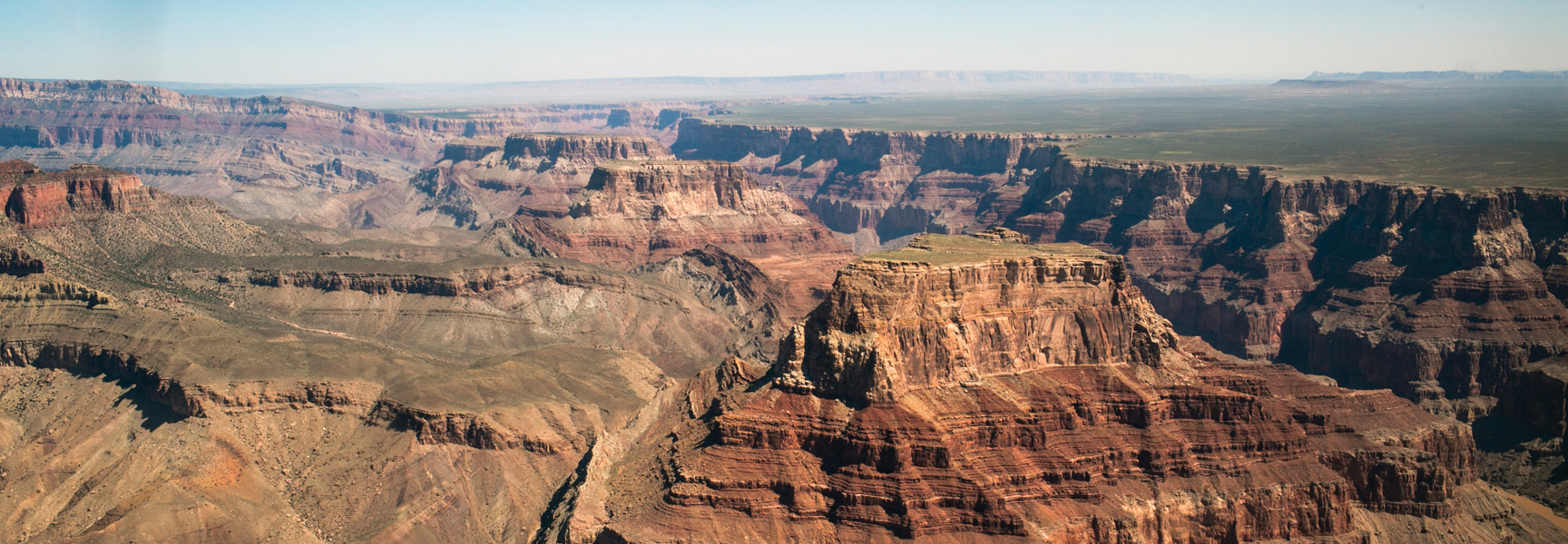 A sweeping aerial view of the Grand Canyon in Arizona shows its vast, layered red rock buttes and canyons under a clear blue sky.