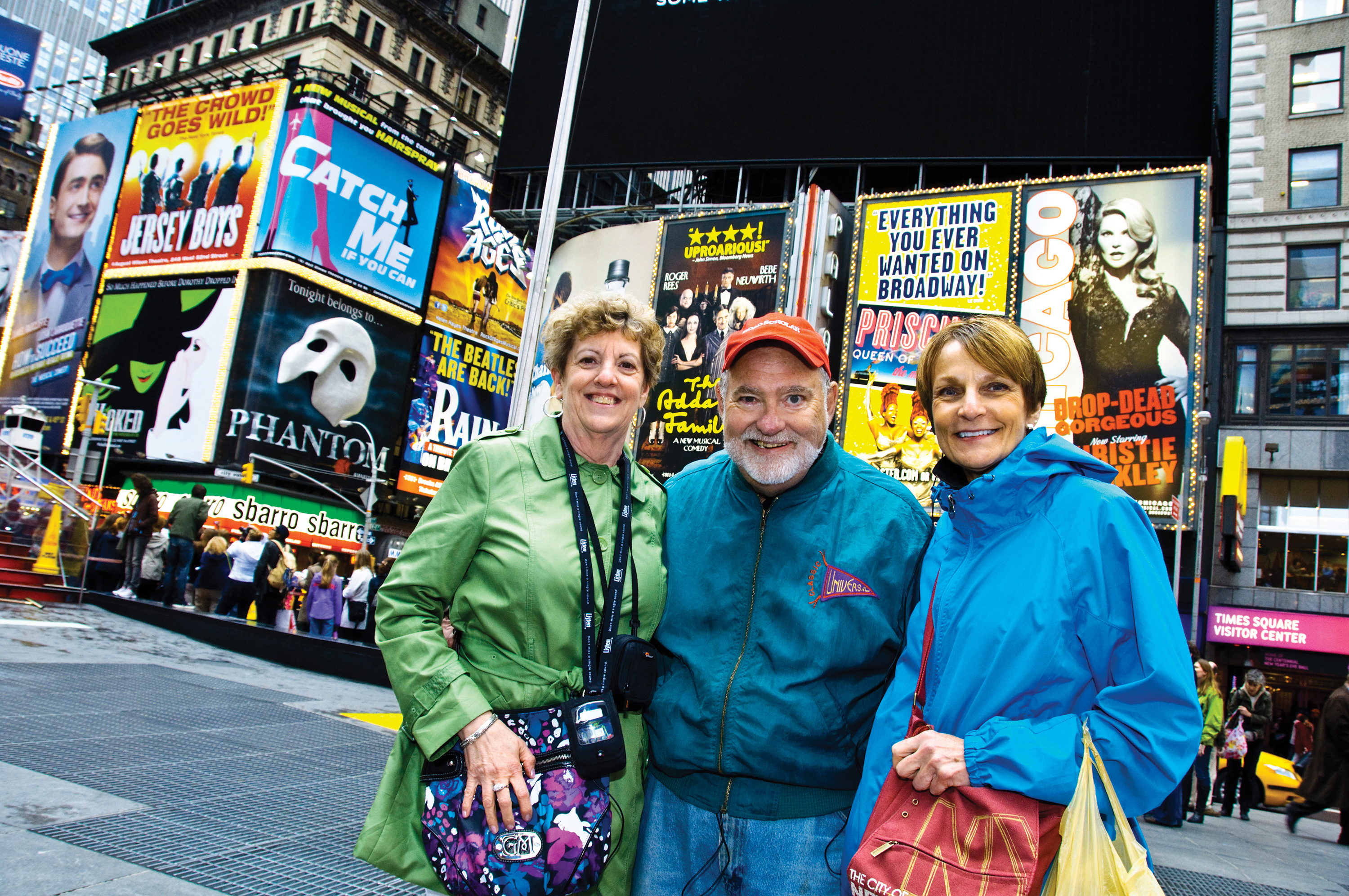 Three people smiling in Times Square, New York, with vibrant Broadway theater billboards in the background.