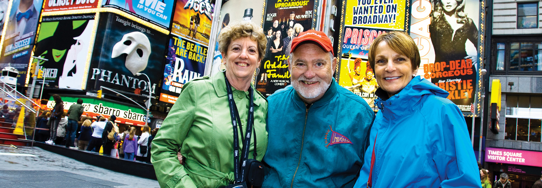 Three smiling adults pose for a photo in New York's Times Square, with numerous bright Broadway show billboards behind them.