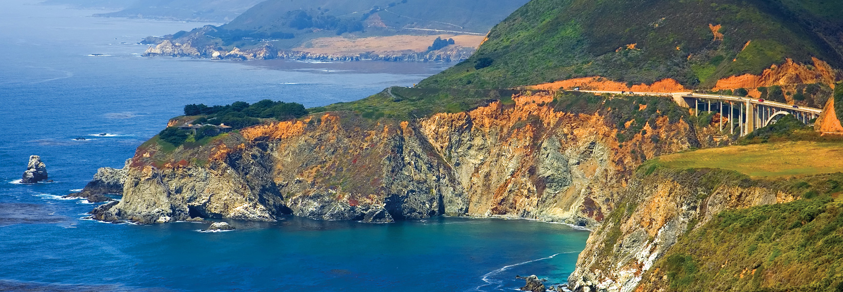 A dramatic view of the rugged California coastline in Monterey, with a highway bridge spanning a cliffside over the bright blue Pacific Ocean.