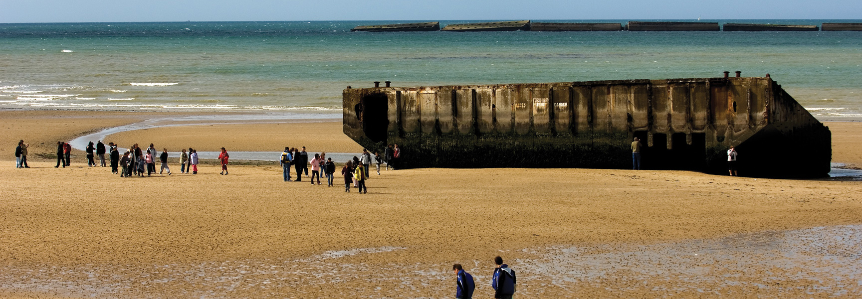 People walk on a sandy beach in France, viewing large concrete remnants of the D-Day landings in the sand and sea.