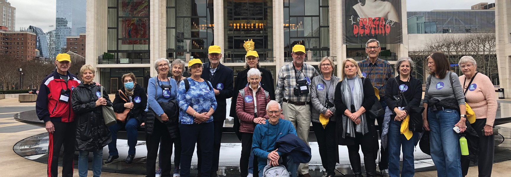 A large group of adults on an opera tour smile for a photo outside the Metropolitan Opera House in New York.