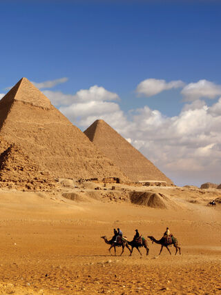 People ride camels across the desert landscape in front of the iconic Great Pyramids of Giza in Egypt.