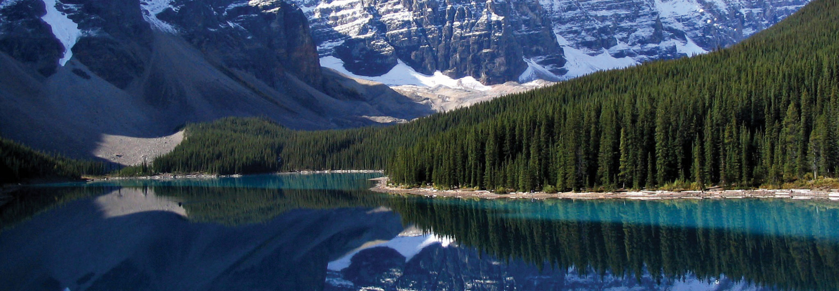 A turquoise lake in Alberta reflects the snow-capped Rocky Mountains and a dense pine forest lining its shores.