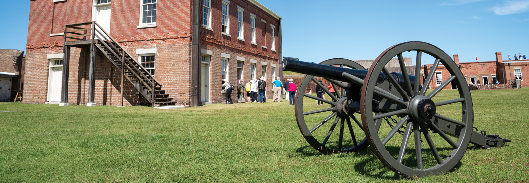 A large cannon sits on a grassy field before a historic brick fort, with a group of tourists gathered near the building.