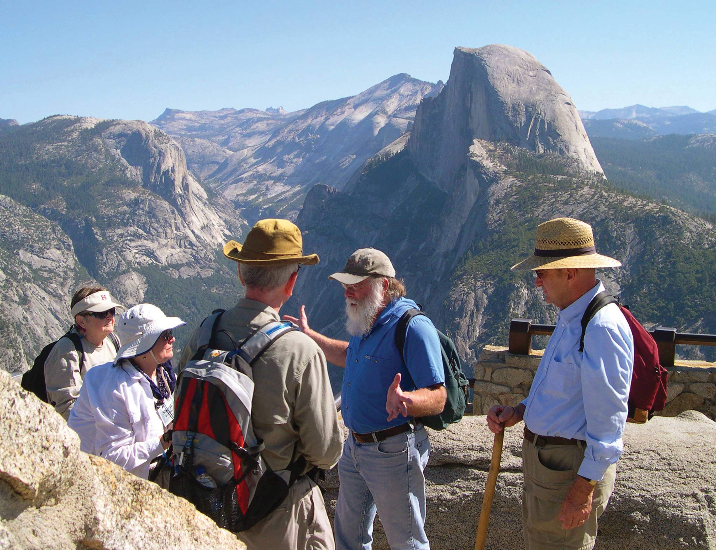A group of hikers listens to an expert guide at a scenic overlook featuring Half Dome in Yosemite National Park, California.