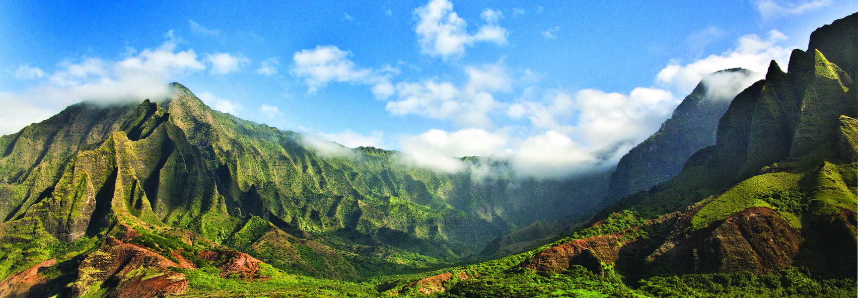 Lush, jagged green mountains of Kauai in Hawaii overlook the vibrant blue ocean under a clear sky with soft white clouds.