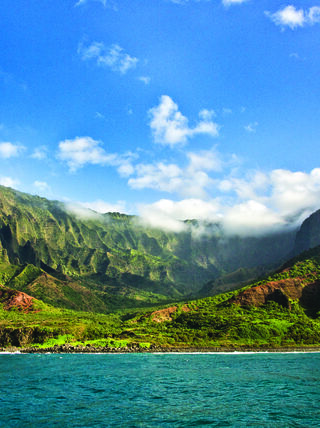 Lush, jagged green mountains of Kauai in Hawaii overlook the vibrant blue ocean under a clear sky with soft white clouds.