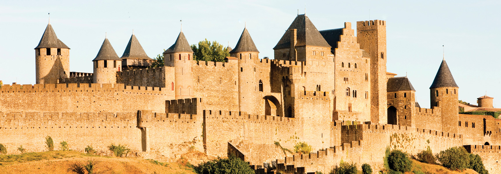 A sprawling stone castle with numerous towers and fortified walls sits on a grassy hill in France, illuminated by golden sunlight.