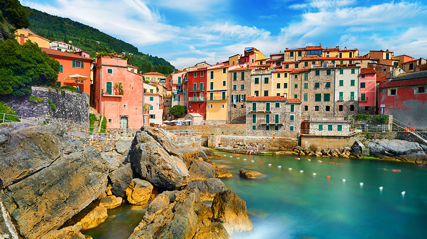 Colorful buildings of Tellaro, Italy, line a rocky cove filled with turquoise water under a partly cloudy sky.