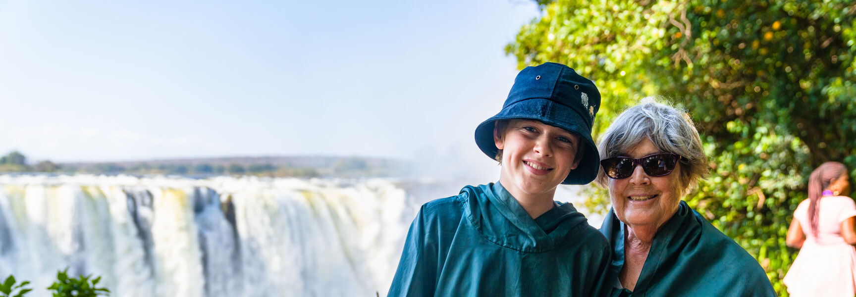 A grandmother and her grandchild wearing green ponchos pose in front of the cascading Victoria Falls during an Africa safari.