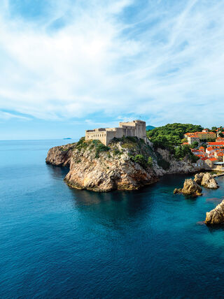 A medieval stone fortress overlooking the majestic Adriatic Sea and a historic coastal city with red-roofed buildings in the Balkans.