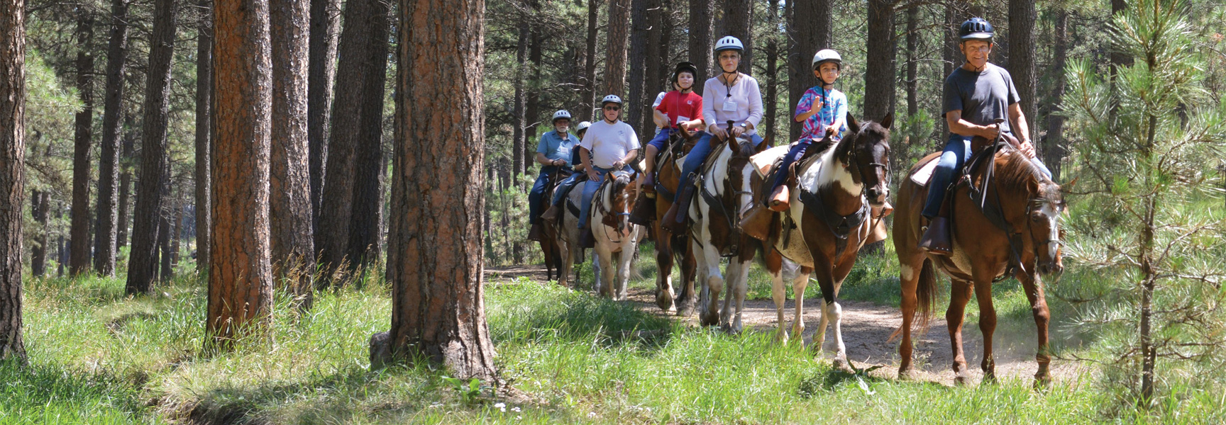 A group of grandparents and grandchildren ride horses on a trail through a pine forest in South Dakota.