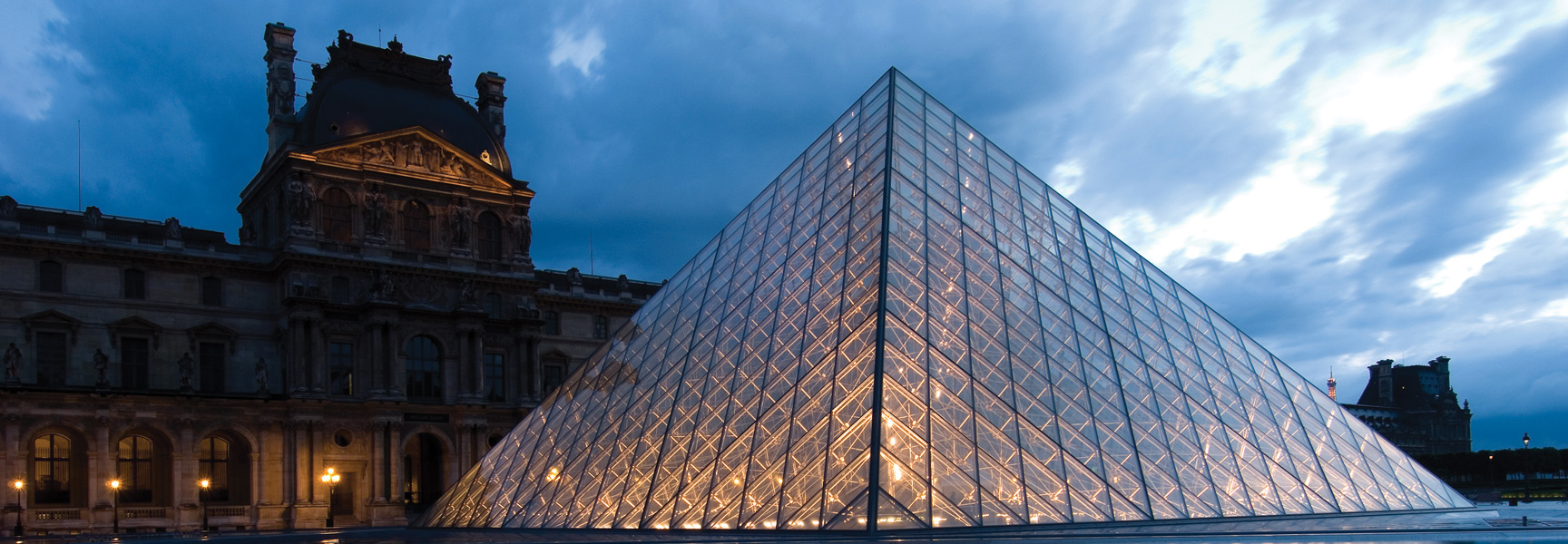 The illuminated glass pyramid of the Louvre Museum in Paris, France, stands against a dramatic cloudy sky at dusk.