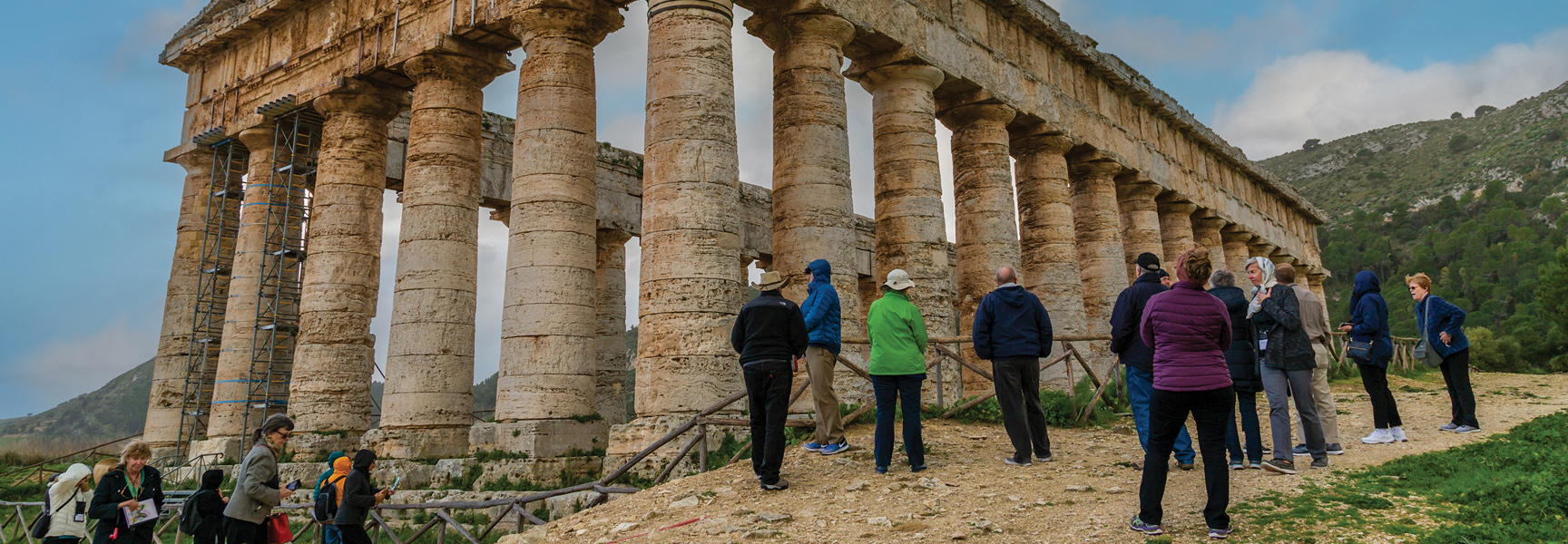 A group of travelers marvels at the ancient Greek temple of Segesta with its large stone columns on a green hillside in Sicily, Italy.