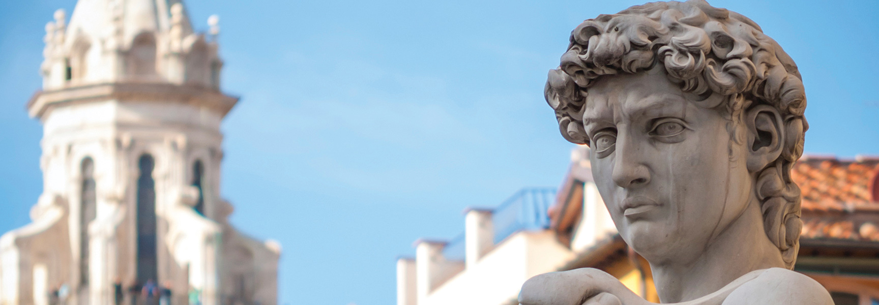A close-up of Michelangelo's famous David statue in Florence, Italy, with a historic tower blurred in the background against a bright blue sky.