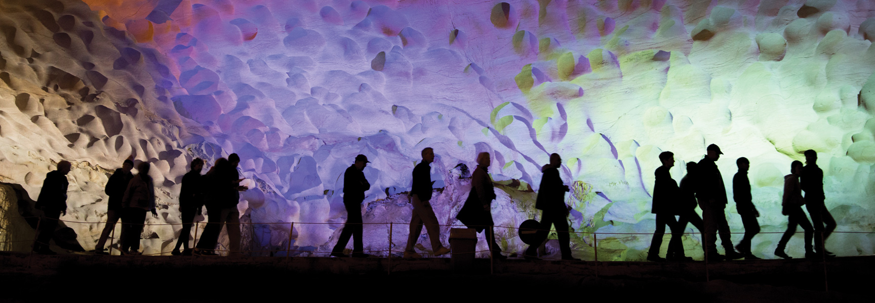 Silhouettes of people walk along a pathway through a dramatically lit cave with purple and green lights in Vietnam or Cambodia.