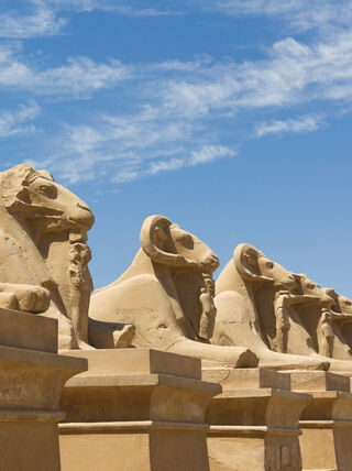 A row of ancient ram-headed sphinxes stands on stone pedestals at a historic site in Egypt under a bright blue sky with wispy clouds.