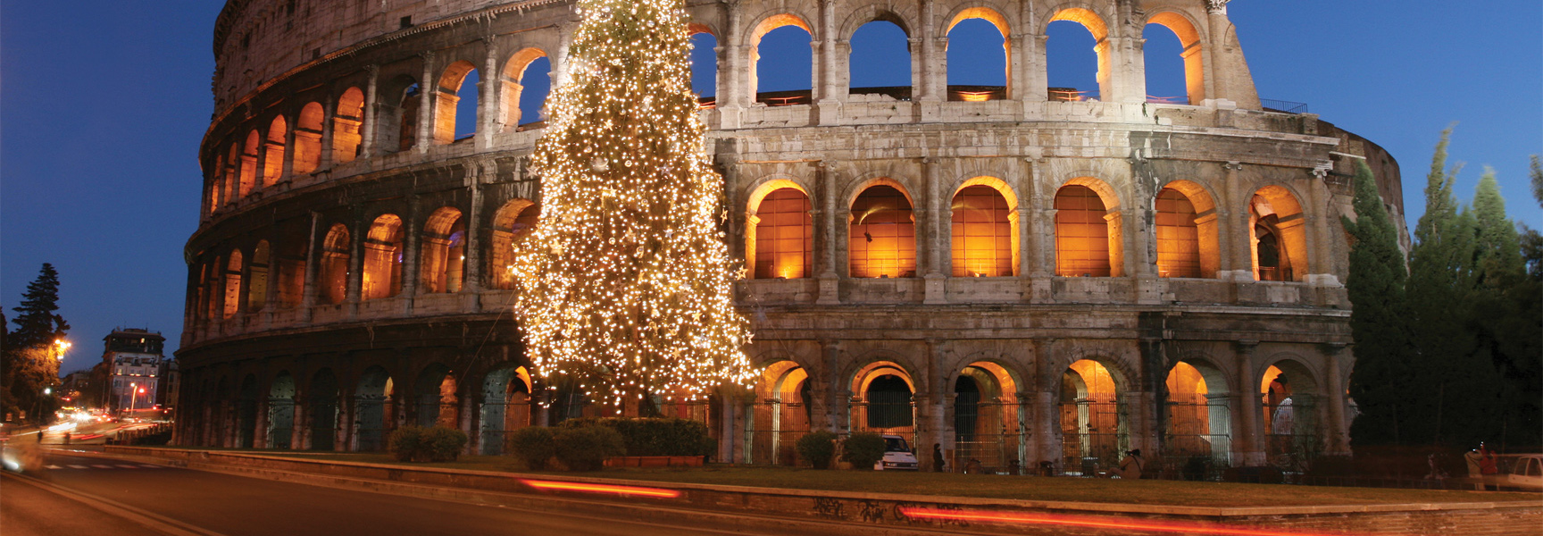A large, lit Christmas tree stands in front of the illuminated Colosseum at night in Rome, Italy.