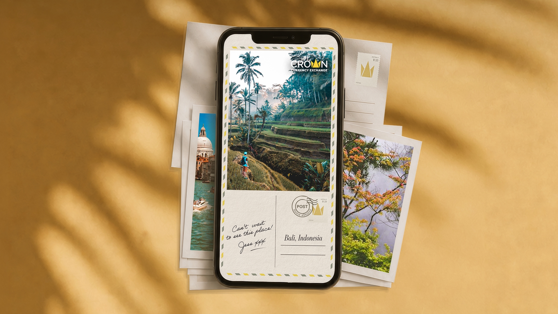Smartphone displaying a Crown Currency Exchange postcard ad styled as a vintage airmail postcard, showing a Bali rice terrace scene with a hiker, a handwritten note reading "Can't wait to see this place! Jess xxx," and the caption "Bali, Indonesia." Travel photo prints of a European canal and autumn foliage are scattered behind it on a warm golden background.