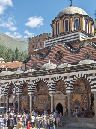 Crowds of people visit the historic Rila Monastery in Bulgaria, featuring its iconic black and white striped arches and ornate frescoes against a mountainous backdrop.