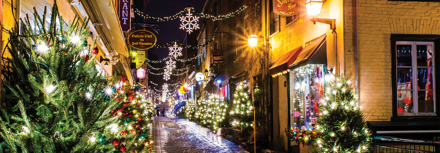 A festive cobblestone street in Québec is brightly illuminated at night with Christmas trees and hanging snowflake lights for the holidays.