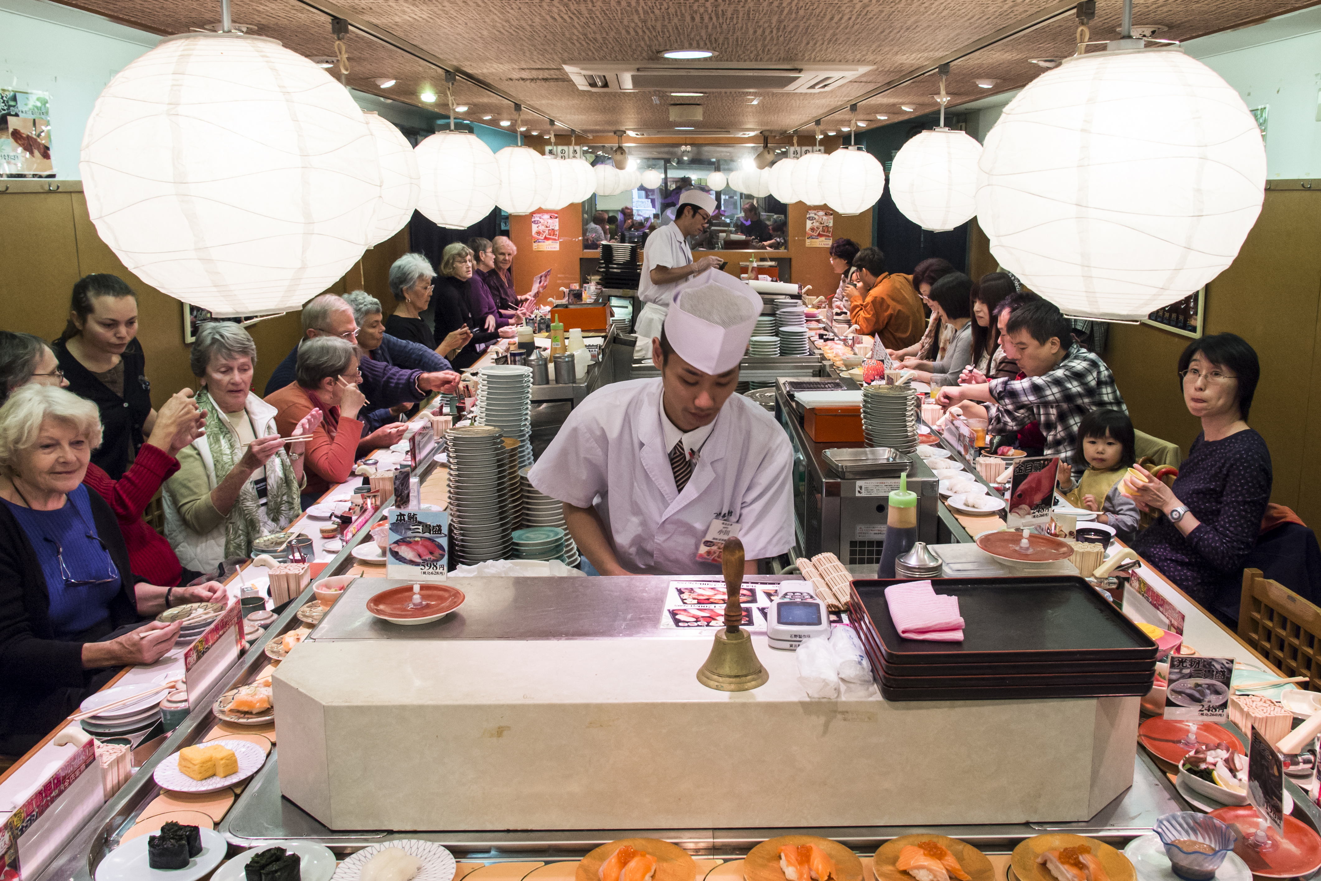Diners sit along a conveyor belt at a sushi restaurant in Japan as chefs prepare food under large, spherical white paper lanterns.