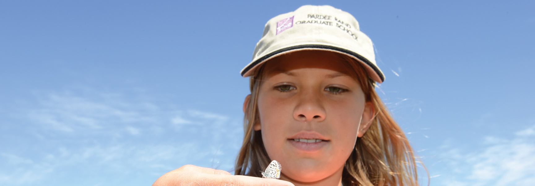 A young girl wearing a baseball cap looks at a small butterfly on her finger under a bright blue sky in Washington.