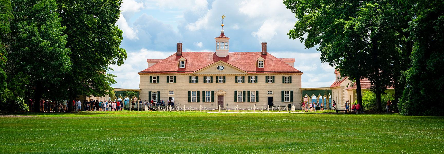 The historic Mount Vernon mansion in Virginia is viewed from across a vast green lawn, with tourists gathered near the entrance.