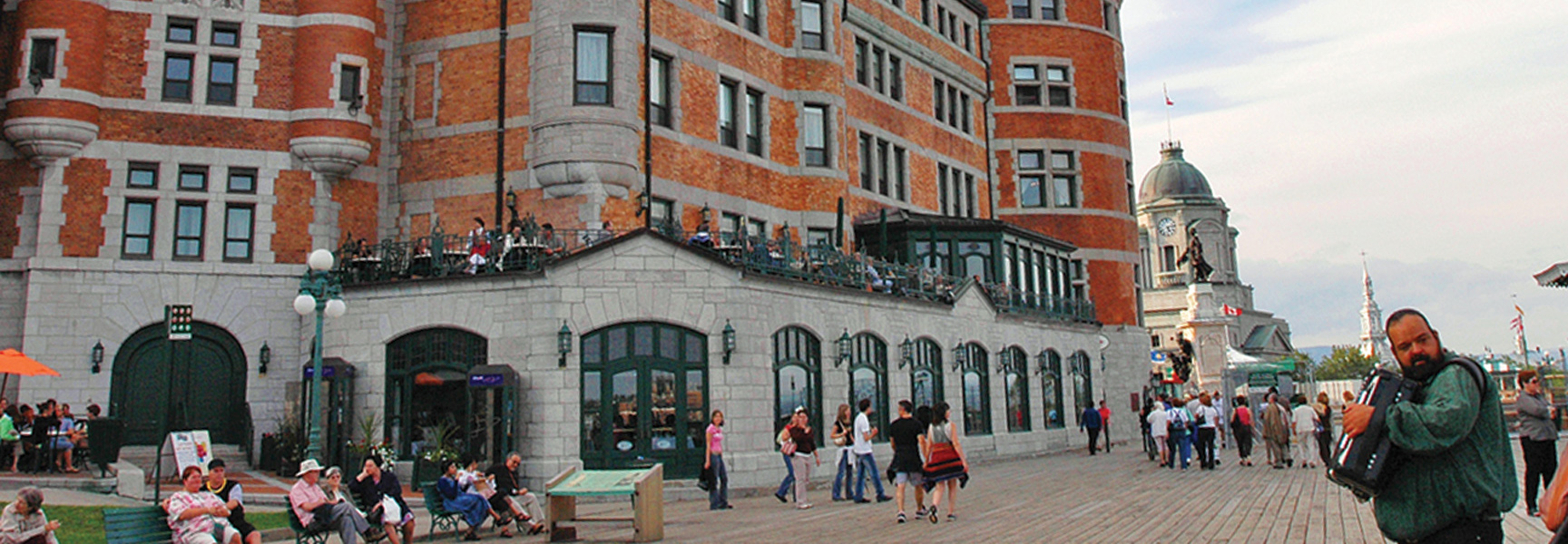 A street musician plays the accordion for people on a boardwalk in Québec, with a large, historic brick and stone building behind them.