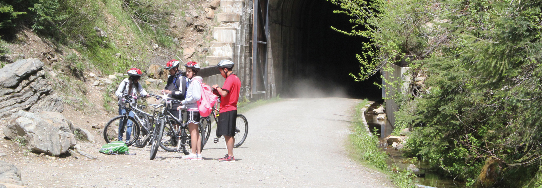 A group of bikers wearing helmets take a break on a dirt trail in Idaho before entering a dark tunnel.