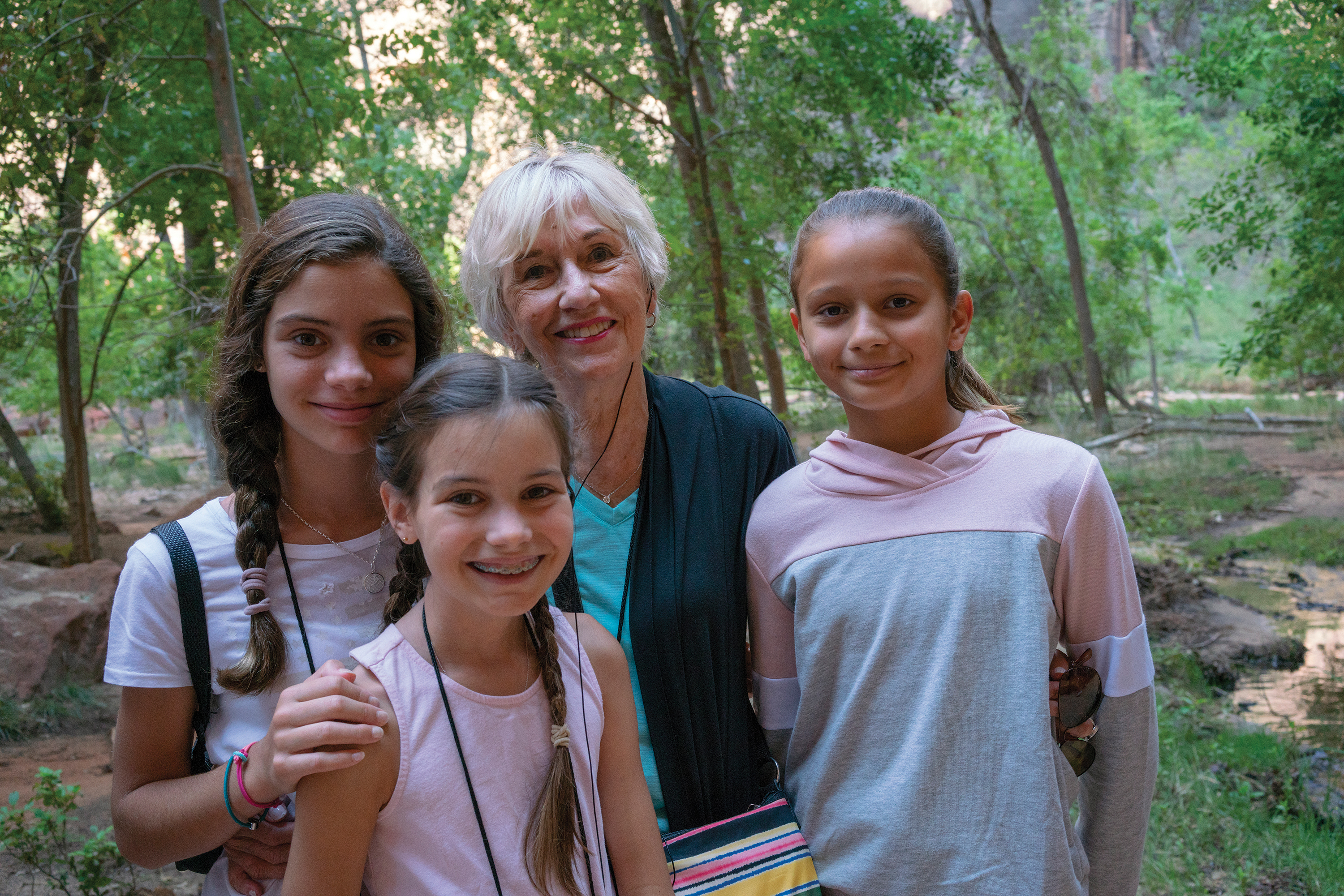 A grandmother and three young granddaughters smile together in a lush, wooded area during a family trip to Zion National Park.