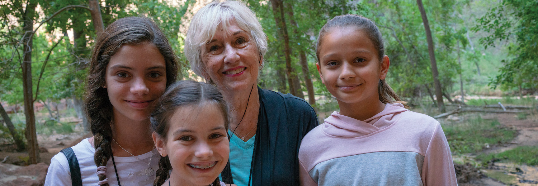A grandmother and her three granddaughters smile together outdoors in a lush green forest in Zion National Park.