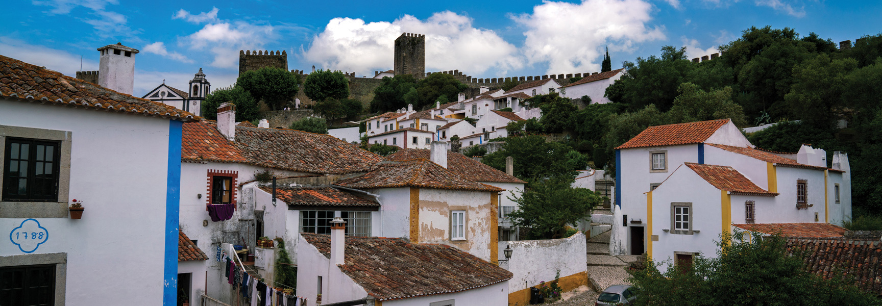 A scenic view of a historic hillside village in Portugal, with white-washed houses, red tile roofs, and a castle on the hill.