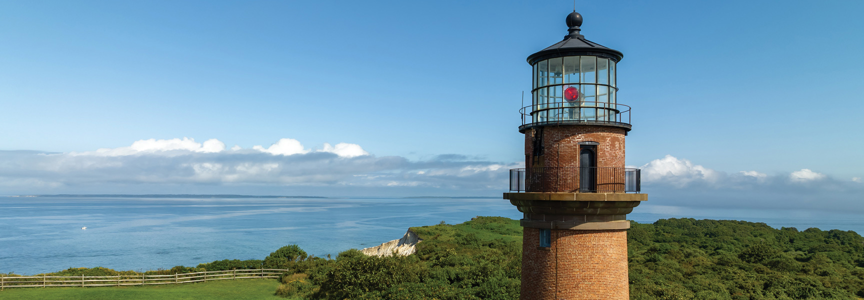 A brick lighthouse on a green cliff in Massachusetts overlooks the blue ocean under a clear sky.