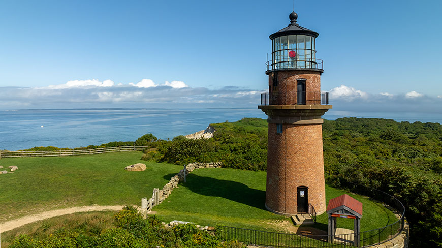 The red brick Gay Head Lighthouse in Aquinnah, Massachusetts, stands on a grassy cliff overlooking the ocean under a blue sky.