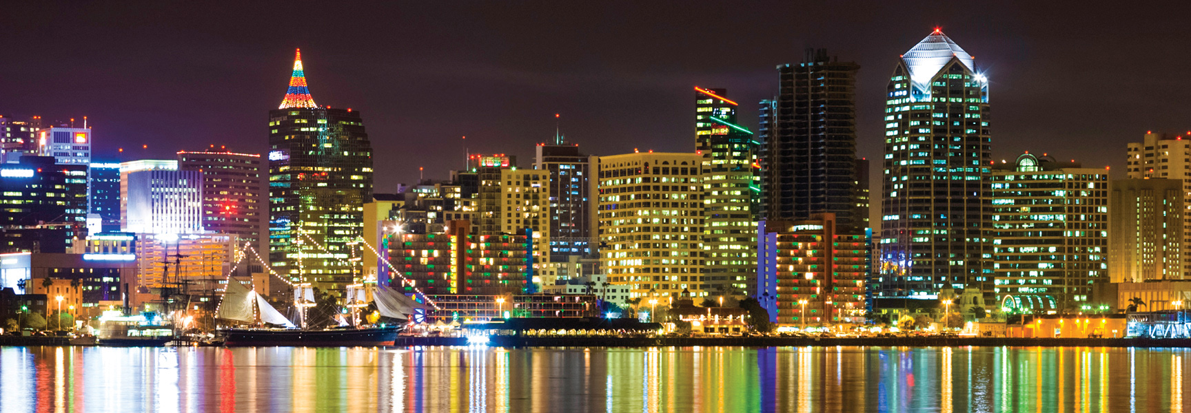 The San Diego, California skyline is illuminated with festive holiday lights at night, with a tall ship sailing in the bay in the foreground.