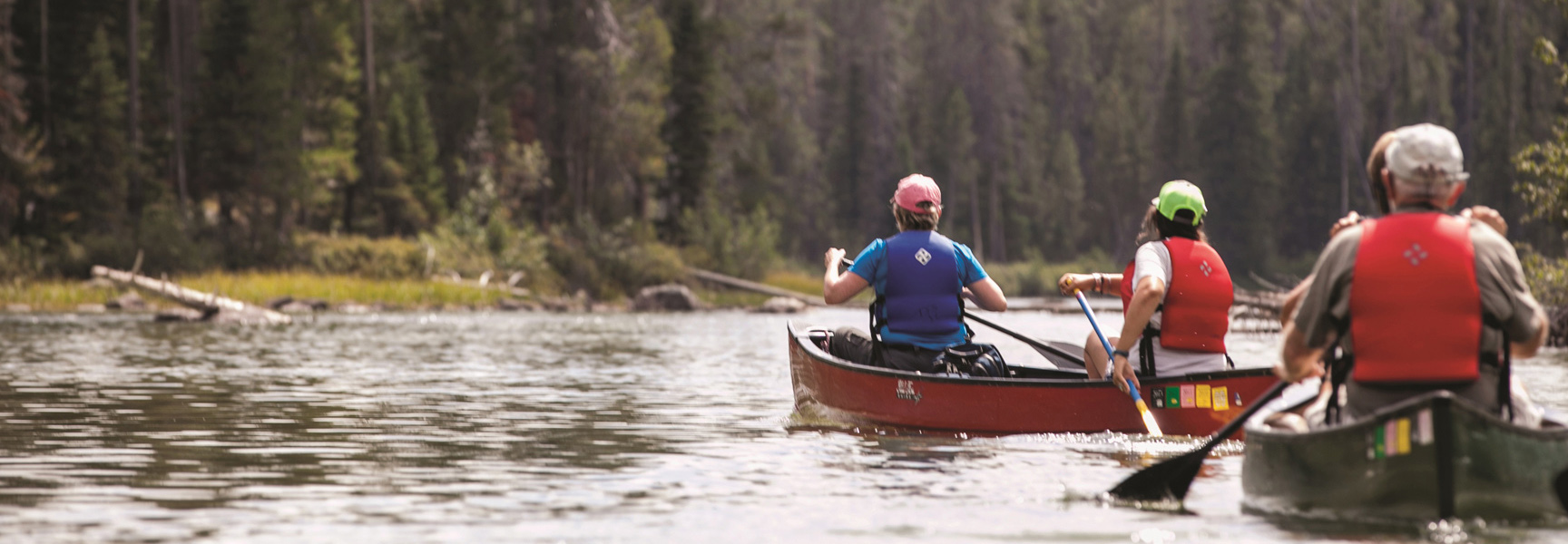 A group of people paddle in red and green canoes on a calm river in Wyoming, with a dense forest on the distant shore.