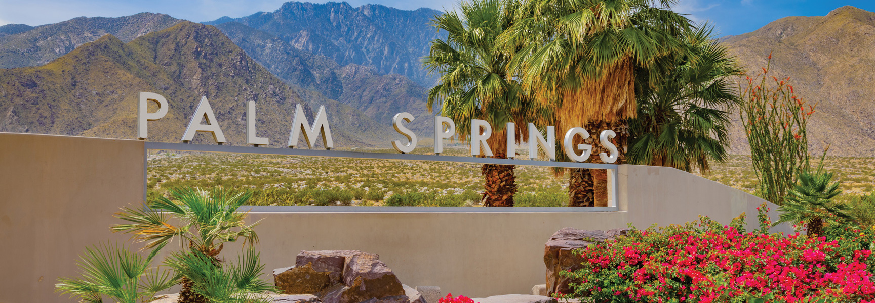 The Palm Springs welcome sign with palm trees and flowers in the foreground and the San Jacinto mountains in the background in California.