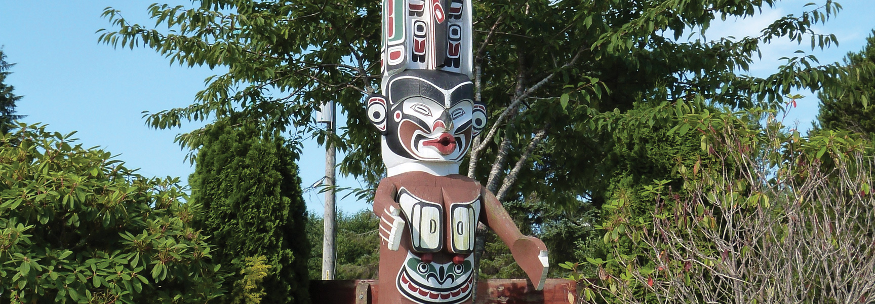 A colorful, carved wooden totem pole stands amongst lush green trees under a blue sky in Western Canada.