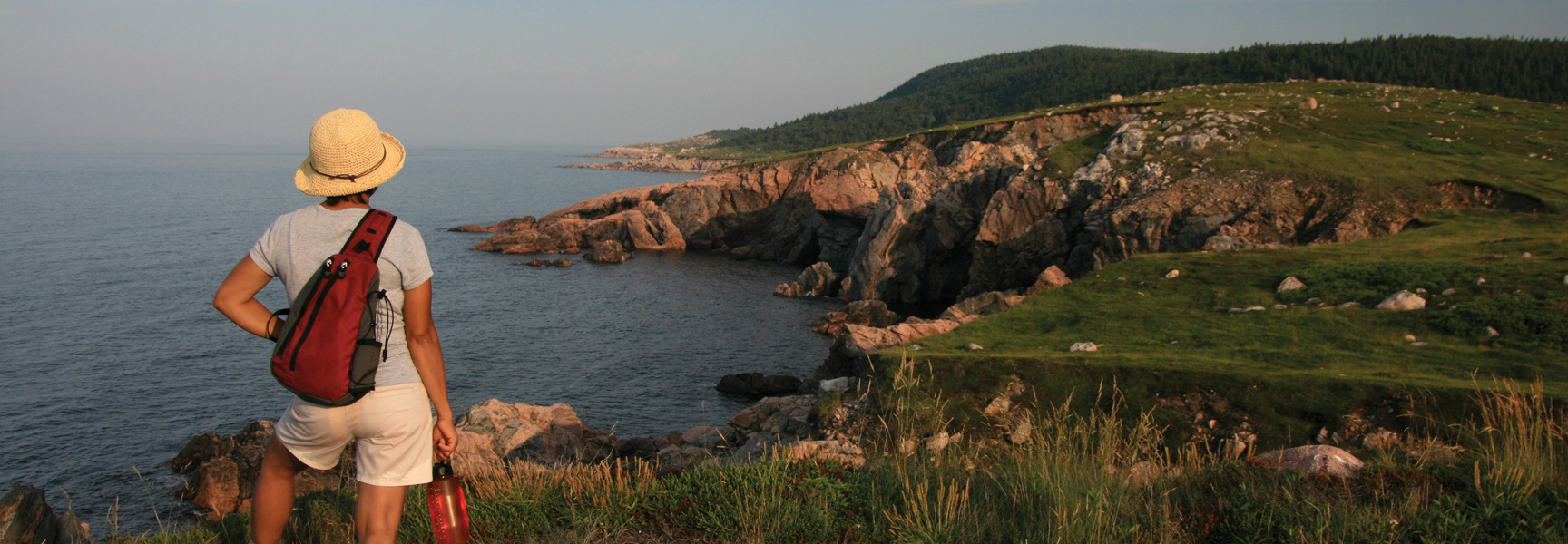 A hiker wearing a hat looks out over the rocky coastline of Cape Breton Highlands National Park in Nova Scotia at sunset.