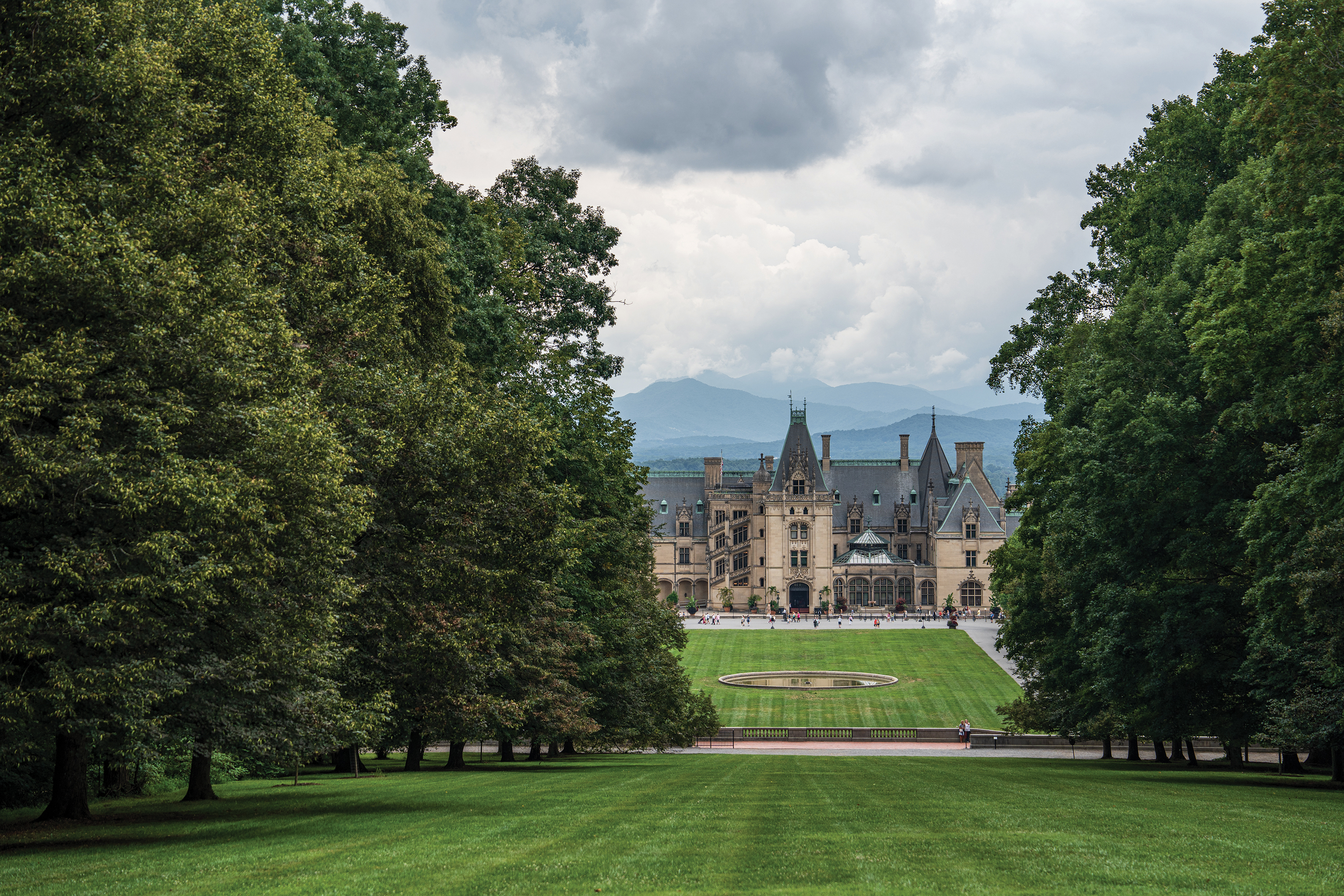 The historic Biltmore Estate in Asheville, North Carolina, framed by lush green trees with a vast lawn and mountain peaks in the distance.