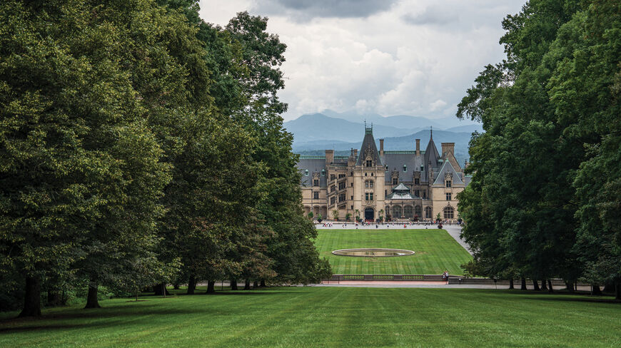 The historic Biltmore Estate in Asheville, North Carolina, framed by lush green trees with a vast lawn and mountain peaks in the distance.